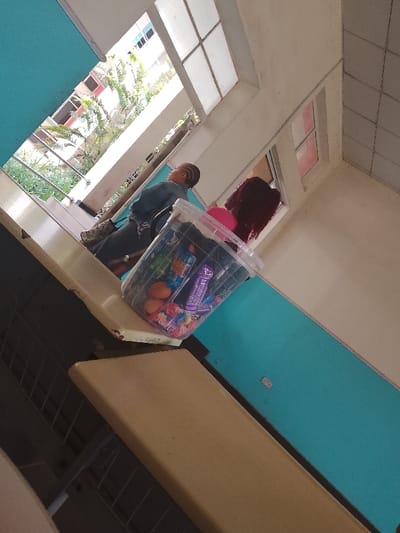 A medium-shot, high-angle photograph depicts an indoor scene. Two individuals, one with reddish-brown hair, are visible in the background by a window. One person has short hair styled in braids. In the foreground, a clear plastic container filled with various snack items, including eggs, candy, and chocolate bars, rests on a surface. The context of a city is inferred by the prompt and implied through the appearance of an urban setting outside the window. The location is Umueshi, Nigeria.