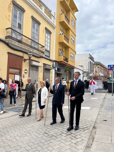 The image shows a procession of four men in suits walking down a narrow street in Las Palmas de Gran Canaria, Spain. They are holding staffs topped with gold finials. The men are walking on a cobblestone street between buildings. The buildings are painted yellow and have balconies. There are other people in the background, some are standing, some are walking, and some are standing on a balcony. The procession is walking towards a woman in a white robe and red sash standing in the distance. The image was taken on a cloudy day. The coordinates (latitude: 28.099, longitude: -15.418) place the location in the historic district of Vegueta, near the Catedral de Santa Ana.
