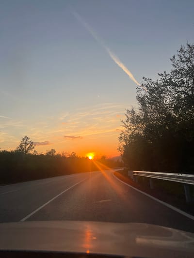The image depicts a sunset over a road in Balkantsi, Bulgaria. The sun is setting directly ahead, creating a bright glare along the road's surface. A guardrail is visible along the right side of the road, bordered by trees. The sky is a mix of orange and blue hues, with contrails present. Part of a vehicle is visible in the foreground on the right side of the image.