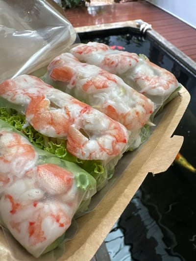 Four fresh spring rolls, filled with shrimp and lettuce, are arranged in a brown cardboard takeout container. The container is placed on the edge of a dark pond, where two koi fish are visible swimming. The pond is surrounded by wooden decking. The overall scene suggests an outdoor setting, likely in a residential or restaurant area with a water feature. The location is specified as Ban Kanchanok 23, Thailand.
