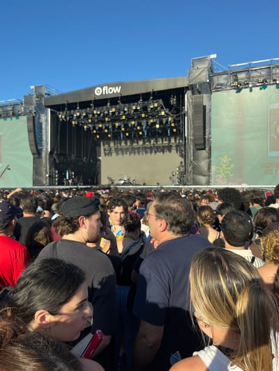 An outdoor concert or festival scene is depicted in San Isidro, Argentina, under a clear blue sky. A large stage structure dominates the background, featuring a prominent "flow" logo on its canopy and multiple arrays of stage lights. Large video screens flank the stage, displaying graphic elements including a green cartoon figure and partial Spanish text that reads "DISPONIBLE LOS Y". The foreground and midground are densely populated by a large crowd of individuals, primarily adults, most oriented towards the stage. No specific interactions are clearly visible within the compact gathering. Identifiable persons in the closer proximity include a male wearing a black cap and t-shirt, seen in profile, and another male with glasses and a dark blue t-shirt, viewed from behind. A person with curly hair and a colorful lanyard is also visible, along with a female with blonde hair in a ponytail in the right foreground. The environment suggests a daytime event due to the bright, natural lighting.