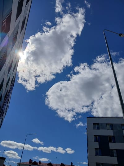 The image captures a bright, sunny day in Gubkinsky, Russia, with a clear blue sky dotted with fluffy white clouds. The perspective is from a low angle, looking upwards. On the left side of the frame, a modern building with a dark, textured facade rises diagonally. The sun, partially obscured by clouds, is visible to the upper left, casting a lens flare that creates a rainbow effect on the building. To the right, a tall, slender street lamp with a light fixture at its top stands against the sky. In the lower right corner, the upper floors of another building with a gray and white patterned facade are visible. Below that, the red-tiled roofs of what appear to be residential buildings can be seen. Several other street lamps are visible in the distance, along with more clouds. The scene depicts a typical urban environment on a pleasant day.