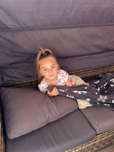 The image shows a young girl lying on a dark-colored outdoor sectional sofa. She is partially covered by a dark gray blanket with white snowflake patterns. The girl has blonde hair tied in a ponytail and is wearing a light-colored pajama set with heart patterns. She is looking directly at the camera with a gentle expression. The sofa is upholstered in a dark fabric and has thick cushions. The background shows a wicker weave pattern, likely part of the sofa's frame or surrounding furniture. The lighting suggests it is daytime, possibly outdoors given the furniture style. The girl is holding onto the blanket, seemingly relaxed or playing. There is no visible text in the image. The location is specified as Waldbröl, Germany.