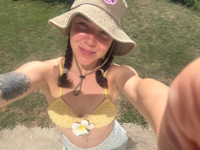 A young adult female with braided brown hair, wearing a beige bucket hat and a yellow bikini top adorned with a white flower, takes a selfie outdoors. She has a tattoo on her upper left arm and a smaller tattoo on her abdomen. The background consists of green grass and a paved surface, suggesting a park or backyard setting. The lighting indicates it is daytime, with sunlight casting shadows. The location is identified as Tvarditsa, Bulgaria.