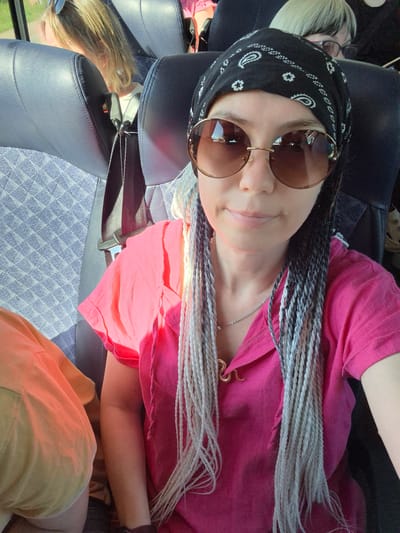 A medium shot shows a woman with braided, multi-colored hair (black, white, and gray) wearing a black bandana with a white paisley pattern, large round sunglasses with brown lenses, and a bright pink collared shirt. She is seated on a bus, as indicated by the dark blue patterned upholstery of the seat and a seatbelt visible. Another person, partially visible in an orange shirt, is seated to her left. The woman is looking towards the camera and smiling slightly. The environment appears to be daytime, with sunlight illuminating the scene. The image does not provide any information about the location being Красный Ключ, Russia.