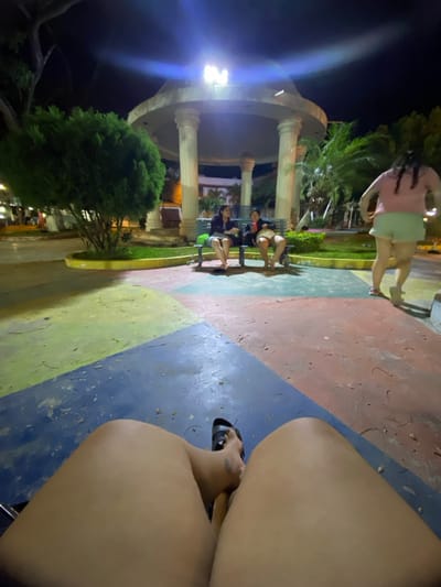 This image captures a nighttime scene within a public park or plaza in General Villamil Playas, Ecuador. The perspective is from a seated position, with the lower legs and right foot of the photographer visible in the immediate foreground; the right foot wears a dark sandal. The ground consists of large, interlocking geometric shapes in blue, red, yellow, and green.
In the midground, two individuals are seated on a park bench positioned in front of a white, multi-columned gazebo. The person on the left wears a dark top and light shorts, holding a beverage. The person on the right wears a light top and dark shorts, holding an object, and appears to be interacting with the individual to their left. To the right of the bench, a third individual, dressed in a light pink top and light green shorts, is in motion, facing away from the camera and moving towards the right side of the frame.
The background is dominated by the brightly illuminated gazebo, which features an overhead light causing a significant lens flare. Various trees and bushes, including a dense green shrub on the left and palm trees on the right, are visible. Distant buildings and artificial lighting are faintly discernible under a dark night sky.