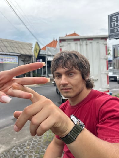 A horizontally oriented, close-up shot depicts a fair-skinned man in a red t-shirt holding his right hand up, which wears a silver watch with a green patterned band on his left wrist. His shoulder is visible on the right. His face is tilted towards the camera. In front of him, a second hand with long, light-pink manicured nails is making a "peace sign." The background indicates an outdoor setting with buildings and street signs visible in Munggu, Indonesia. A white truck with red stripes is partially visible behind the man's head. The sky is overcast.