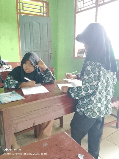 An indoor scene in what appears to be a classroom in Wanguk, Indonesia, captured on November 26, 2025, at 15:04. The walls are light green. Two female individuals, likely students, are present.
The individual on the left is seated at a dark red-brown wooden desk. She wears a black hijab and a long-sleeved shirt with a green and white geometric pattern. She holds a white pen in her right hand, which rests on an open notebook containing written text. Her left hand is positioned near her temple. A light blue book with an illustration lies on the desk to her left. She is looking towards the camera with a neutral expression.
The individual on the right is standing or leaning next to a desk, partially obscured by bright sunlight from a window. She also wears a black hijab and a matching green and white geometric patterned long-sleeved shirt. Her hands are positioned over an open notebook or paper on the desk, with her right hand holding a pen or pencil. Her face is not clearly visible.
In the background, a light blue wooden door with vertical panels is visible on the left wall, next to a wooden-framed window with horizontal grates. On the right wall, two wooden-framed windows with vertical grates allow bright sunlight into the room. A small, fragmented white object is visible on a desk in the immediate foreground, bottom right.