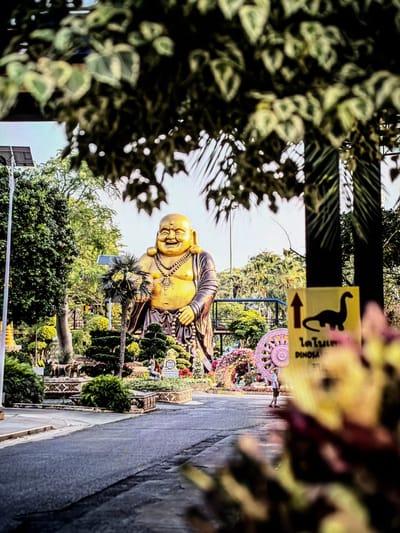 In the city of Vientiane Capital, Laos, a wide, paved pathway leads to a large, golden statue of a smiling Buddha. The statue, adorned with a golden necklace, stands prominently amidst a lush garden filled with manicured trees, ornate planters, and colorful flowers. To the right of the Buddha, a yellow sign with a silhouette of a dinosaur and Thai text is visible. In the background, various trees and foliage create a vibrant green backdrop under a bright sky. A solitary figure, a person wearing a white shirt and dark shorts, walks away from the viewer on the pathway, approaching a decorative floral arch and a large, circular pink structure. The foreground is partially obscured by blurred yellow and green foliage.