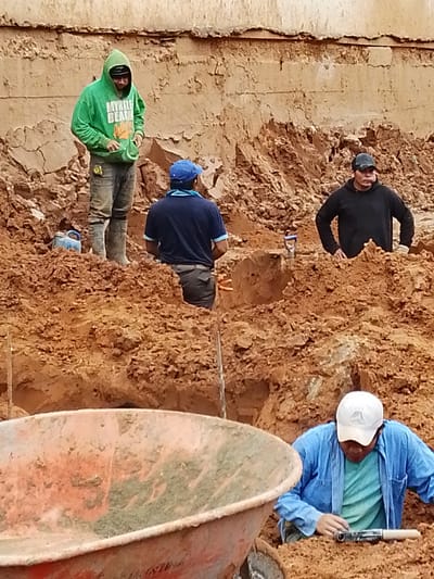The image depicts four individuals engaged in what appears to be construction or excavation work in Santa Cruz de la Sierra, Bolivia. The environment is characterized by a large pile of reddish-brown dirt and a concrete or earthen wall in the background.

In the foreground, a person wearing a white cap and a blue long-sleeved shirt over a turquoise t-shirt is bending down, appearing to be examining or working with a tool. To the left, the edge of a large, orange wheelbarrow is visible.

Further back, another person in a black hoodie and a dark cap is partially visible, standing amidst the dirt. To the left of this individual, another person in a dark blue t-shirt and dark pants is also standing.

Dominating the left side of the image is a person wearing a bright green hooded sweatshirt with "MYRTLE BEACH" printed on it, and dirty gray pants. This individual is also standing in the dirt. A small blue container or bucket is visible near their feet. The ground is uneven, with piles and trenches of soil. The overall scene suggests an outdoor work site with ongoing labor.