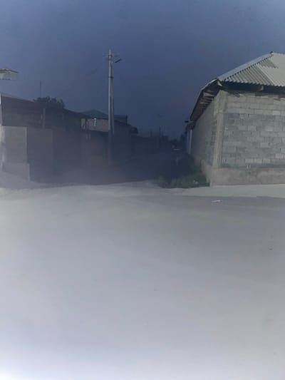This media file depicts an outdoor scene, likely in Боғистон, Tajikistan. The perspective is from a low angle, looking upwards and to the right, with the image rotated such that the horizon is vertical.
The primary subjects are a building and a road. The building appears to be constructed of concrete blocks with a corrugated metal roof. It is situated on the left side of the frame. The road runs along the right side of the building and stretches into the distance. The road surface appears to be asphalt or a similar material, and it is somewhat hazy or dusty.
There are some objects visible. A utility pole with wires is present in the middle-left of the image, appearing to be part of the infrastructure along the road. In the distance, along the road, a small group of colorful objects, possibly people or items, are visible. The overall scene suggests a somewhat rural or developing area.
The time of day appears to be daytime, possibly late afternoon or early morning, given the soft lighting and shadows. The weather seems to be clear, although the haze on the road might indicate dusty conditions. There are no discernible emotions expressed in the image. No visible text is present. The orientation of the image suggests it was taken while moving, possibly from a vehicle.