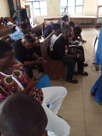 The image shows a group of people sitting on wooden benches in a room in Yenagoa, Nigeria, located at coordinates 4.931, 6.268. The individuals are sitting in a semi-circular formation. Several are looking down at their phones. Most of the people are wearing casual clothing. The walls are off-white, and the floor is tiled. The benches are wooden and are placed in a row. There is a blue bench in the foreground. There is an electrical cord leading from an electrical outlet on the wall to a device in the center of the floor. The image is taken from a low angle.