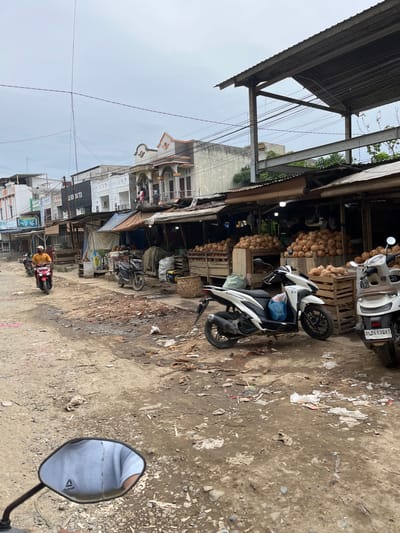 In the city of Kota Kuala Simpang, Indonesia, a dirt road lined with market stalls is captured under an overcast sky. The stalls, constructed with corrugated metal roofing and wooden supports, display large quantities of coconuts piled high in wooden crates. Several motorcycles are parked in the area, including a red scooter with a rider in a yellow shirt, a white scooter, and a dark scooter. The ground is uneven and littered with debris, including plastic bags, paper, and dirt. Buildings with various architectural styles and signage, such as "SORAYA TEX" and "The Great Barbershop," are visible behind the market stalls. The scene depicts a bustling, albeit somewhat unkempt, market environment.
