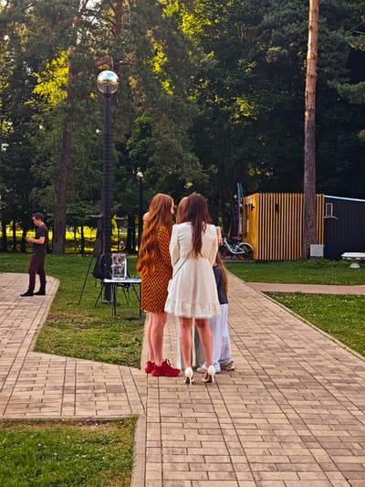 The image captures an outdoor scene in Svistukha (Kuzyayevskiy S/O), Russia, likely a park or recreational area. A paved brick pathway, interspersed with grassy areas, forms the foreground and leads into the background. Several people are visible. On the left, a man in a dark t-shirt and maroon pants stands looking at his phone. In the center, a group of three women are engaged in conversation. One woman on the left has long, reddish-orange hair and wears a patterned orange and black dress with red shoes. Next to her, a woman in a light-colored dress with white heels stands facing away from the camera. A third woman, partially visible, stands behind her. In the background, a tall lamp post with a spherical light fixture stands prominently. Beyond the people and pathway, trees and foliage form a dense green backdrop. A wooden structure with vertical slats and a dark-colored section is visible to the right, with a bicycle parked nearby. The overall atmosphere suggests a casual gathering or event.