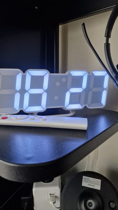 This is a close-up, slightly angled shot of a white digital clock with blue LED display, showing the time "08:08". The clock is mounted on a dark surface, likely a desk or shelf. To the right of the clock, a white remote control with red buttons is leaning against the same surface. In the background, a black security camera is visible, with a label that includes the text "Lomoo", "Shenzhen Belong Technology Co., Ltd.", "CE ROHS", and an email address "remetyeah.net". A white power adapter is plugged into an outlet behind the camera. The scene is indoors, and the lighting appears to be from the digital clock itself, casting a blue glow. The overall impression is of a functional, modern home or office setup.