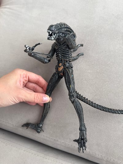 An overhead medium shot shows a person's left hand holding the leg of a black, detailed alien action figure. The alien figure is standing on a textured gray surface, possibly a couch. The figure is dark gray with some metallic bronze and blue accents visible on its body. Its mouth is open, revealing sharp teeth and an inner jaw. The alien has a long, segmented tail that curves to the right, and two pipe-like appendages protrude from its upper back. The hand is supporting the figure by its upper thigh. The background is a plain, textured gray fabric. The location is identified as Nizhnyayarkeyevo, Russia.