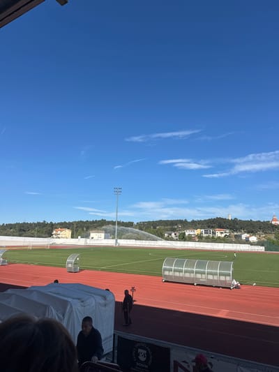 Captured from an elevated perspective, the scene showcases an athletic field in Valpaços, Portugal, under a bright blue sky with wispy clouds. The green field is flanked by a red running track. A sprinkler system is in operation, irrigating the field. On the far side of the field, houses dot a hillside. In the foreground, a white tent-like structure partially obscures the view. People are visible near the tent. A sign bearing the words "Friends and Wine SONIM" is also in view.