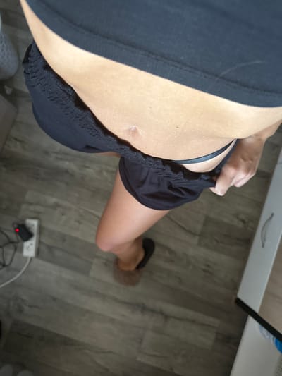The image shows a person's midsection from a high-angle perspective, focusing on their abdomen and upper thighs. The person is wearing a black sports bra and black shorts that have been pulled down slightly, revealing a portion of a black thong. A small, light-colored scar is visible on the person's abdomen. The floor is made of grey wood-look laminate. In the background, a white electrical outlet with a red-lit plug is visible on the floor. To the right, a white surface, possibly furniture, is partially visible. The image does not contain any information that would allow for the determination of its location in Basarabeasca, Moldova.