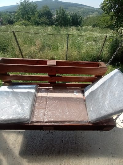 The image shows a wooden pallet swing seat with three cushions placed on it. The swing is suspended by chains, visible on the right side of the image, and appears to be outdoors with a background of lush green vegetation and rolling hills. The cushions are covered in plastic protective wrapping, with one on the left and one on the right, and another in the center. The wooden structure of the swing seat is made from horizontal planks. The ground beneath the swing appears to be a concrete surface. The scene is set in a natural environment, likely a backyard or a garden, with trees and a fence visible in the background. The location is specified as Bârnova, Romania.