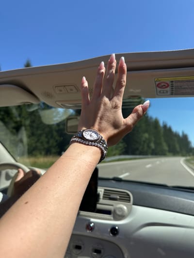 A person's arm is extended out of a white convertible car, with their hand reaching towards the sky. The arm is adorned with a diamond bracelet and a watch. The car is driving on a road surrounded by trees under a clear blue sky. The image appears to be taken in Vöhrenbach, Germany, on a sunny day.