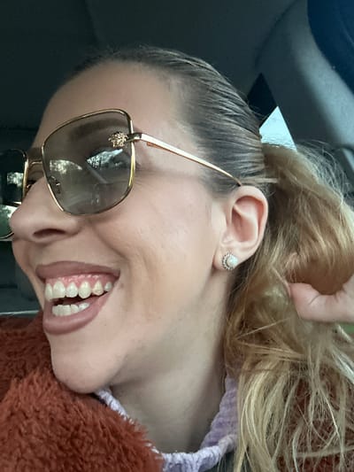 A woman is smiling, with her head tilted. She is wearing gold-framed sunglasses with a visible Versace emblem. Her hair is styled in a bun. She has a pearl-like stud earring in her left ear. She is wearing a fuzzy brown garment. The image was taken indoors, possibly inside a car. No information exists to suggest that the image was taken in Basildon, United Kingdom.