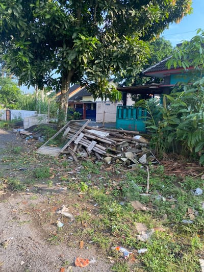 The scene is a dirt road in front of a residential building in Kecamatan Piyungan, Indonesia (latitude: -7.827, longitude: 110.482). A large tree with green leaves stands in the foreground. A pile of wooden debris is strewn on the ground next to the tree. The building is a two-story structure with a blue roof. A blue metal gate stands in front of the building. There is lush green vegetation growing around the area. There is no one visible in the image. The debris includes several long wooden planks and other assorted pieces of lumber. The ground is covered with dirt and debris, including leaves, twigs, and plastic trash. The environment is a residential area, likely a quiet neighborhood.