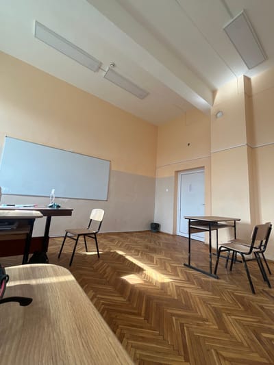 This image depicts an unoccupied classroom or educational space in Voysil, Bulgaria. The room features a two-tone wall scheme, with the upper sections painted light peach and the lower sections a lighter, off-white or beige, showing signs of wear and discoloration. The ceiling is white with visible structural beams and multiple rectangular LED light fixtures, along with a circular smoke detector. The floor is covered in a light to medium-toned wooden parquet, laid in a herringbone pattern. Sunlight streams into the room, casting distinct rectangular bright patches onto the floor.

Furniture includes three desks and at least three chairs. In the foreground, a light-wood-topped desk is partially visible on the left. Further back on the left, another desk with a dark top supports a clear pump bottle (likely hand sanitizer) and other small, indistinct objects, with a light-wood-and-black-metal chair positioned in front of it. On the right side of the room, a third desk with a light wooden top and a black metal frame is present, accompanied by two light-wood-and-black-metal chairs, one placed next to the desk and another stacked behind it. A large, rectangular white board is mounted on the left wall. A closed white door with some text affixed to it is visible on the right wall, near a black trash can positioned in the corner. No individuals are present in the scene.
