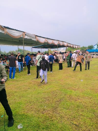 The scene takes place in Karangmalang, Indonesia, at the coordinates -7.47 latitude and 111.064 longitude. It features a large gathering of people under a series of white canopies with brown trim. The ground is covered in green grass. The people are casually dressed, some are standing around, others are talking, and a few are walking. There are a variety of objects in the scene including a few plastic chairs, a discarded plastic cup, and a small, abandoned black rope lying on the ground. The canopies are supported by metal poles. The sky is overcast. A few trees are visible in the background. A building is also visible in the background, though it's unclear what type of building it is.