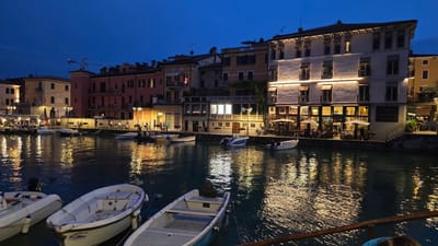 This is a twilight shot of a picturesque canal in Peschiera del Garda, Italy. The scene is illuminated by the warm glow of streetlights and interior lights from buildings lining the canal. Several small boats are moored in the water, their reflections shimmering on the surface. People are seated at outdoor tables at restaurants and cafes along the waterfront, enjoying the evening.

The buildings are a mix of architectural styles, with pastel-colored facades and balconies. One building prominently displays a sign that reads "Hotel Bell Arrivo". The sky is a deep blue, indicating that night has fallen, or is about to. The overall mood is peaceful and inviting, suggesting a relaxing evening in a charming European town. The water appears calm, with gentle ripples reflecting the lights.