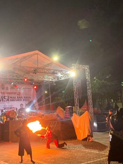 The scene takes place at night in Kecamatan Batu, Indonesia, at the coordinates -7.867, 112.514. There is a stage set up outdoors, with a large white canopy overhead and a metal truss structure holding up lighting fixtures. The stage has a large red sign that says "MALANG RAYA MSI AWARD 2025." There are several people on and around the stage, including a man playing a drum set, a man with a microphone, and a person in a black suit performing a fire-breathing act. Two people dressed in white sheets are standing to the right of the stage. The stage is set up in a paved area with a few trees and other buildings in the background. The scene is illuminated by stage lights and several other streetlights. The sky is dark with a few stars visible. The atmosphere is festive and celebratory.