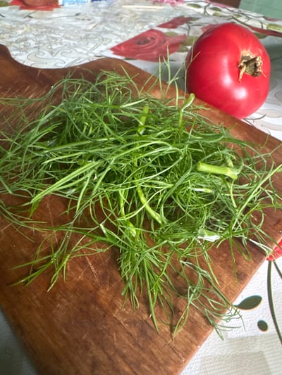 The image shows a wooden cutting board with a pile of fresh dill on it. To the right of the dill, there is a ripe red tomato. The cutting board is placed on a patterned tablecloth, which has floral designs in red and white. The scene appears to be in a kitchen or dining area. The image does not provide any information about the city of Красное, Russia, or any specific location within it. There are no people visible in the image.