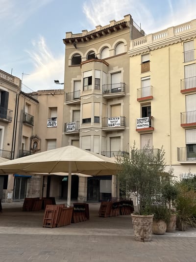 The image depicts a street scene in Igualada, Spain, likely a public square. In the foreground, several large, folded cream-colored umbrellas are set up, covering an outdoor seating area that appears to be associated with a cafe or restaurant. Piled neatly beside the seating area are numerous stacked wooden chairs.
Behind the umbrellas, a row of multi-story buildings lines the street. The architecture features a mix of styles, with balconies, arched windows, and ornate decorative elements. Several banners are visible hanging from the balconies of these buildings. One banner, in Catalan, reads "DRESCANSAR ES UN DRET" which translates to "Resting is a right". Another banner, also in Catalan, states "MENYS TAULES MÉS PLACA" meaning "Fewer tables, more square". A third banner reads "DISCOTECA MUNICIPAL", indicating a municipal disco or nightclub.
A small niche in one of the buildings contains a statue of a religious figure. The sky above is a pale blue with wispy white clouds, suggesting it might be late afternoon or early morning. The ground is paved with cobblestones. The overall impression is of a quiet urban environment, possibly before the start of business hours or during a less busy period. No people are visible in the image.