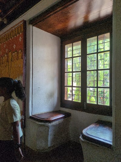 The image depicts the interior of a historical building, identified as Mattancherry Palace in Kochi, India. On the left, a person, seen from the mid-torso up and facing away from the camera, stands near a wall featuring a large, intricate mural. The individual has dark hair tied in a ponytail and wears a light-colored, short-sleeved top and dark lower garments. The mural, rendered in earth tones including gold, red, and white, portrays multiple figures in a traditional Indian artistic style.
To the right, a deep window recess is illuminated by natural light entering through a large, multi-paned wooden window. The window consists of two vertical sections, each divided into multiple smaller rectangular panes by a dark grid frame. Green foliage is visible beyond the window, suggesting an outdoor garden or trees. Within the window recess, two built-in stone benches are present: one to the left under the window, and another extending from the right wall into the recess. Both benches are topped with dark, polished wooden cushions featuring rounded edges and decorative trim. The walls are thick and painted an off-white or light beige. A dark wooden beam runs horizontally across the top of the frame, above the mural and window. The floor is dark and unadorned.