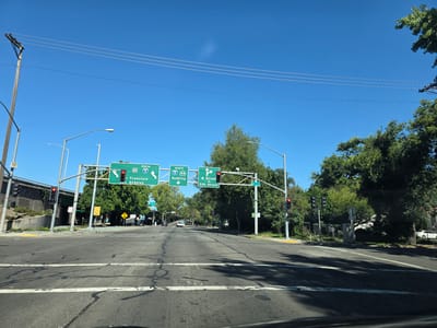 The image captures a sunny daytime view of a multi-lane road in Sacramento, California. The scene is dominated by overhead highway signs directing traffic.
On the left, a sign indicates "SOUTH" on Interstate 80 and Interstate 5, with arrows pointing towards "San Francisco" and "Los Angeles." To the right of this, another sign shows "NORTH" on Interstate 5 and State Route 99, with an arrow for "Redding." Further to the right, a sign directs traffic to "W Street OR 5th Street." Traffic lights are visible, with some showing red.
The road itself is paved and marked with white lines. Lush green trees line the sides of the road, and the sky is a clear, bright blue. Telephone poles and power lines are present. In the distance, a white car is visible. The overall impression is that of an urban or suburban intersection with clear signage for major routes. The time of day appears to be midday due to the bright sunlight and clear sky.