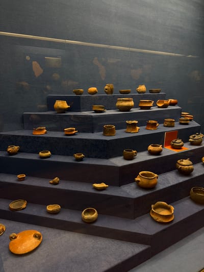The image shows a display of ancient pottery artifacts on a tiered, dark blue structure within a museum. The artifacts are primarily small, earthenware vessels of various shapes and sizes, some appearing to be bowls, pots, and amphorae, with colors ranging from light brown to terracotta. The lighting highlights the textured surfaces of the pottery. The background is a dark, textured wall with a faint horizontal reflection of light. The scene suggests an exhibition focused on historical ceramics, likely in a controlled museum environment. The image does not provide information about the city of Las Palmas De Gran Canaria, Spain.