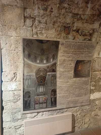 The image shows a sign with a picture and text in Greek and English, mounted on a stone wall. The sign is titled "MIDDLE BYZANTINE MARBLE ICONOSTASI" and describes fragments of an iconostasis from the Monastery of Hosios Loukas in Boeotia, Greece. The image on the sign depicts the interior of a church with an iconostasis, mosaics, and architectural details. The sign provides a numbered list of identified fragments with descriptions. The surrounding environment appears to be an indoor space with rough stone walls and a white heating unit visible at the bottom. The sign and its display suggest an informational exhibit about historical artifacts.