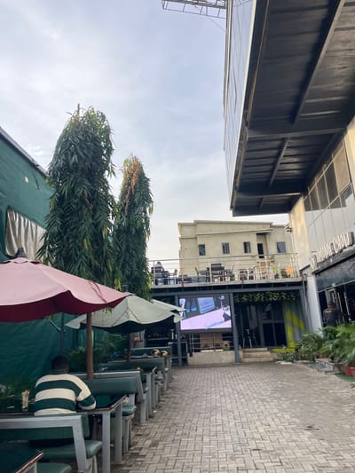 The image depicts an outdoor seating area at a venue in Karum, Nigeria. The scene is set in what appears to be an urban environment, with a modern building visible on the right side of the frame, featuring large windows and a sign that reads "DOMINION CHOW." In the foreground, there are multiple outdoor tables and seating arrangements, some covered by umbrellas, suggesting a restaurant or cafe. Paving stones form the ground surface. Several tall, slender trees are planted in the middle ground, adding greenery to the setting. In the background, another building with multiple windows is visible, along with a balcony area with tables and chairs. A large LED screen is mounted on a structure in the middle of the outdoor space, displaying an image. One person is seated at a table in the foreground, facing away from the camera, wearing a horizontally striped shirt. Another individual is standing on a walkway in the background. The sky is overcast, indicating a daytime setting.