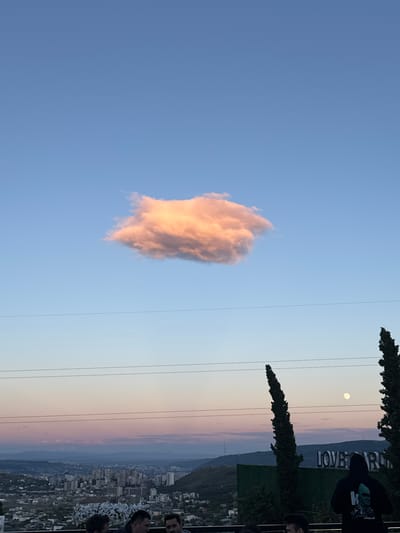 A single, fluffy cloud is illuminated by the setting sun against a clear blue sky. Below, a cityscape stretches out towards a hazy mountain range. A large, white sign with the words "LOVE AR RU" is visible, partially obscured by cypress trees. The moon is also visible in the sky. The image appears to be taken at dusk, with the sky transitioning from blue to soft pinks and oranges. The overall mood is serene and picturesque. The scene is set in Mukhatskaro, Georgia, as indicated by the location context.
