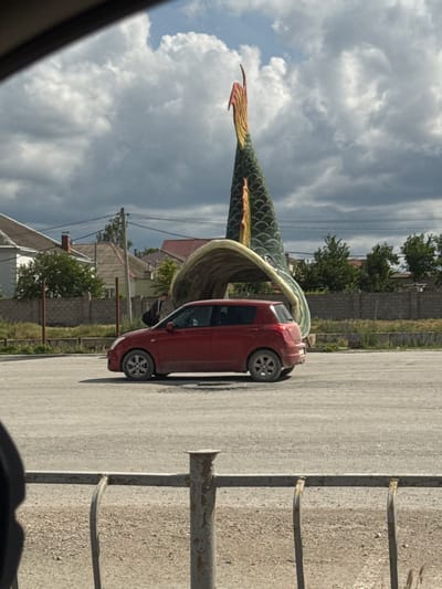 The image, taken in Добровское сельское поселение, Ukraine, depicts a red car parked on an asphalt surface. The car is angled towards the right side of the frame, with its rear wheel clearly visible. In the background, a large, green, fish-like sculpture is prominently featured, its tail extending upwards and its body curving around to partially obscure the car. The sculpture appears to be made of a metallic material with a scaled texture. Beyond the sculpture and car, residential buildings with red roofs are visible, interspersed with trees and a brick wall. The sky above is cloudy and appears to be during the daytime, possibly late afternoon or early evening given the lighting. The overall setting suggests an outdoor location, possibly a public space or a yard. There is no visible text in the image.