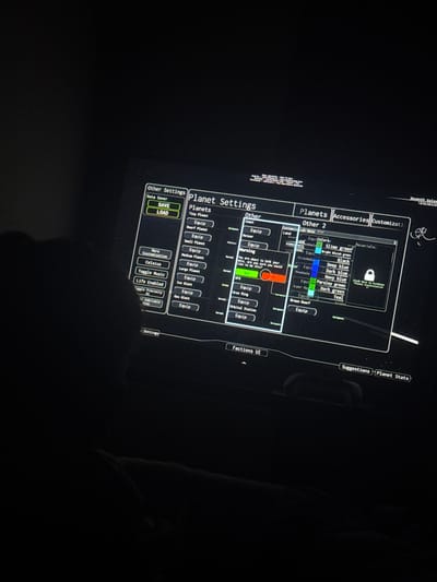 The image shows a screen displaying a game interface, likely a space simulation or planet customization game. The menus visible include "Other Settings," "Planet Settings," "Planets," "Accessories," and "Customizati." There are options to save and load data, equip various planetary features (e.g., comets, meteors, UFOs), and customize planet appearance with different colors and materials. A warning message is displayed asking if the user is sure about saving the data. The environment is dark, indicating low lighting conditions in the room where the picture was taken. The game also has suggestions and planet stats. The information "Seventh Galax 184638438" is visible, potentially indicating a game version or build number. The location of Welshpool, UK is not present in the image.