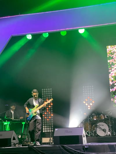 The image shows a concert or live music performance scene, likely indoors. A guitarist, wearing a dark jacket and light shirt, is playing a blue and green bass guitar. Behind the guitarist is a keyboard player who is wearing a white shirt and black suit jacket. Both are illuminated by bright stage lights and a wall of small LED lights. A drum kit is visible in the upper right corner, labeled "Ludwig." The stage is decorated with vertical LED panels. A floral pattern is displayed on a large screen at the top. Loud speakers are placed on the stage. The location is likely the city of Salembaran, Indonesia, but this cannot be determined by information in the image.