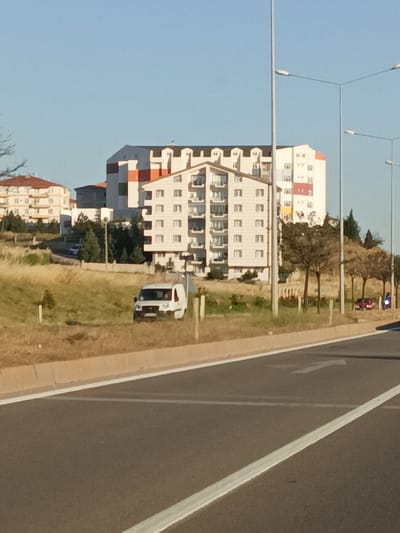 This image depicts a street scene with residential buildings and a road in the foreground. The buildings are modern, with multiple stories and balconies, and some have colorful accents. There are streetlights visible on the right side of the image. In the middle ground, a white van is parked on the side of a grassy area. Behind the van, more buildings and trees are visible. The road in the foreground has white lane markings. Two small cars, one red and one blue, are parked further down the road on the right. The sky is clear and blue, suggesting it is daytime and fair weather. The scene appears to be in a developed, suburban or urban area. No people are clearly visible, though it's possible there are occupants in the van or cars. The overall impression is of a peaceful, everyday setting.