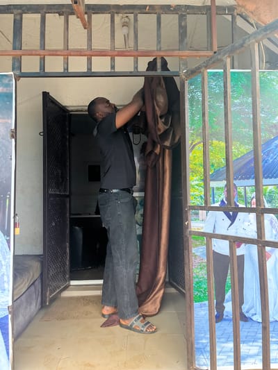 The image shows a man wearing a black t-shirt and dark jeans, with grey sandals on his feet. He is positioned in the center of the frame, reaching up and pulling on a large piece of brown fabric. The fabric appears to be draped over a metal structure, possibly part of a display or a temporary setup.

Behind the man and the fabric, there are large posters visible through a metal grate. These posters depict wedding scenes, with people in formal attire, suggesting the location might be a wedding hall, a photo studio, or a place that displays wedding-related materials. The posters have a light and airy feel, with greenery and white elements.

The setting appears to be outdoors or in an open-air structure, given the lighting and the presence of a metal grate that allows a view of the outside through the posters. The time of day seems to be daytime, as indicated by the bright lighting. There are no explicit cues about the weather, but the light suggests it's not heavily overcast. The overall impression is one of a busy or work-in-progress environment, possibly related to event preparation or display setup. The man's activity suggests he is engaged in setting up or taking down something. No visible text is discernible from the posters. The image is oriented vertically, with the man's feet at the top of the frame.