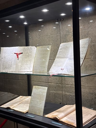 In this media file, ancient manuscripts are displayed within a glass-enclosed exhibit. The scene is lit by overhead lights, creating bright circular reflections on the dark interior surface of the display case, which also reflects the textured stone wall behind the exhibit.

The primary objects are several unfolded manuscripts, appearing as aged parchment covered in dense, handwritten text in a script that is likely Arabic or a related language. One manuscript, positioned at the top of the frame, is spread open and held by clear acrylic panels. Below it, another manuscript is similarly displayed, with a vibrant red ribbon tied around it, suggesting it is a significant artifact. Further down, more pages of text are visible, some appearing as parts of a larger book or scroll. To the right, an open book with yellowed pages and similar script is also on display.

The overall impression is one of historical preservation and academic display. The lighting and reflective surfaces suggest an indoor museum or archive setting. No people are visible, emphasizing the focus on the historical documents. The image captures the quiet reverence of viewing ancient texts, with the surrounding environment designed to protect and showcase these fragile pieces of history. The specific location, Benalmádena, Spain, is mentioned, but no visual cues within the image directly confirm this location beyond it being a display within a Spanish context.
