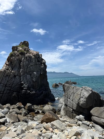 The image shows a rocky coastline with large boulders in the foreground and middle ground. The sea is a vibrant turquoise and a distant mountain range is visible on the horizon under a blue sky with scattered white clouds. The location is identified as Vĩnh Lương, Vietnam. There are no people or animals present in the image. The scene is static and depicts a natural coastal landscape.