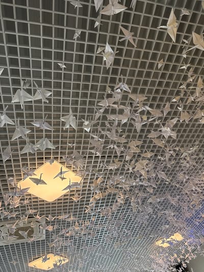 The image shows an indoor art installation. Many white origami cranes are suspended from a white grid structure, creating a visually dense display. The grid structure appears to be attached to a wall or ceiling. Behind the cranes, there are light sources with diamond-shaped covers that illuminate the artwork. The scene is indoors. The image does not show people, subjects, actions, or interactions. The location is Peresvet, Russia.