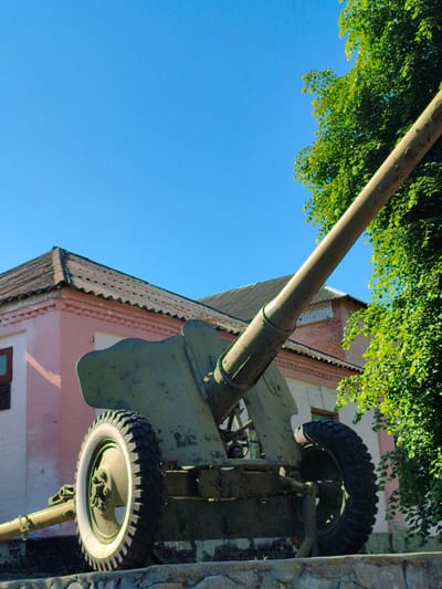 An olive-green military artillery piece, possibly a field gun or howitzer, is the central subject of the image, prominently displayed outdoors under a clear, bright blue sky. The cannon is angled upwards and towards the right, with its long, weathered barrel extending into the upper right corner, partially obscured by green foliage.

The artillery piece itself appears aged, with chipped paint and a rugged texture. It rests on a low, grey stone or concrete base. Two large wheels are visible, equipped with sturdy, dark-treaded tires. The left wheel distinctly shows a white sidewall, a common feature on older vehicle tires.

In the background, a building with a light pink or faded red facade and a dark, corrugated roof is situated. The architecture appears functional and possibly historic, with a small, dark-framed window visible on the left side. Behind the cannon's barrel, another section of the building with exposed red brickwork can be seen. To the right of the cannon, a vibrant green tree or dense bush, lush with leaves, partially frames the scene, illuminated by bright sunlight.

The setting suggests a sunny day with clear weather, given the vivid blue sky and strong illumination. The scene depicts a static display, likely a monument or historical exhibit. No people or visible text are present. Based on the location context, this is likely a memorial or historical piece in Velyka Bahachka, Ukraine.