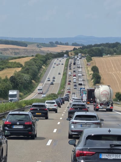 The image shows a multi-lane highway in a rural landscape. The highway is congested with traffic, including cars, trucks, and a tanker trailer. The landscape is characterized by rolling hills with fields of crops and scattered trees. Wind turbines are visible in the distance on a hilltop. The sky is overcast. A black Volkswagen Golf with license plate "PAF FM 86" is in the foreground, to the left. A silver Volkswagen Passat with license plate "GD UX 110" is in the middle ground, to the right. A tanker trailer with the text "NILLEZEN EFFELT" and website "WWW.NILLEZEN.NL" is also visible. The image is captured from a slightly elevated perspective, looking down the highway. Based on visual cues like the road markings, vehicle types, and surrounding landscape, this scene is consistent with a highway in Germany, potentially the A3 autobahn near Dauborn, Germany.