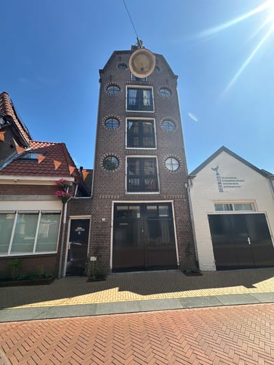 The image shows a street scene in Steenwijk, Netherlands, on a sunny day. The focal point is a tall, narrow brick building with multiple circular windows and a large double door at street level. To the left of this building is a smaller brick building with a red-tiled roof adorned with orange triangular flags, and a large window. To the right of the tall building is a white building with a pitched roof, featuring a large black double door and text on the facade. The foreground is paved with red bricks arranged in a herringbone pattern, with lighter colored paving stones and a dark strip running parallel to the buildings. Shadows from the buildings are cast across the pavement, indicating the time of day. There are no people visible in the image.