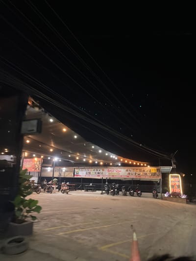 The image captures a nighttime scene outside a restaurant called "Best Kopi & Resto" in Bundar, Indonesia. The restaurant has an outdoor seating area covered by a canopy illuminated by a string of warm lights. Several motorcycles are parked in the paved lot in front of the restaurant. A large sign for "Best Kopi & Resto" is lit up with neon lights. The environment appears to be a commercial area with overhead power lines stretching across the dark sky.